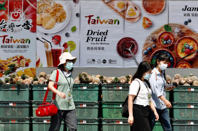 Ananas kasalarının yanından yürüyen insanlar.  Arka duvardaki posterlerde Tayvan'ın tropikal meyvelerinin reklamı yapılıyor
