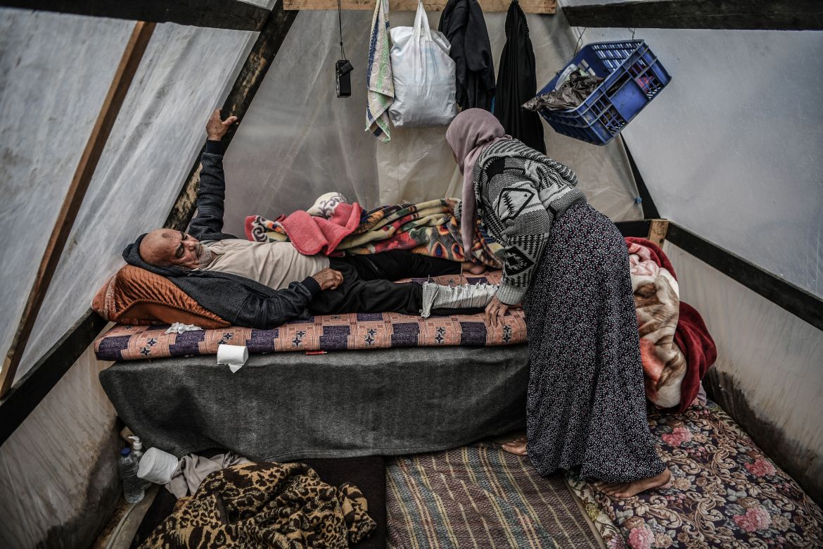 A woman helps a man to lay down on a bed in a makeshift tent during the cold weather as Palestinians, who left their homes, take shelter in the city of Rafah to protect themselves from Israeli bombardment in Rafah