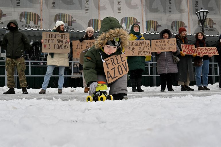 Uma criança brincando na neve com um brinquedo e segurando um cartaz Azov Grátis.  Atrás estão parentes de soldados ucranianos mantidos em cativeiro na Rússia.  Eles também seguram cartazes pedindo a liberdade dos soldados.