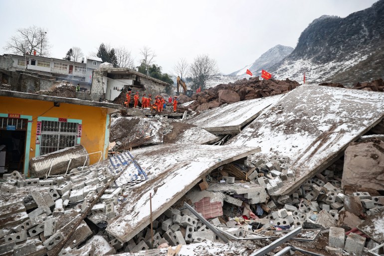 Rescue workers in orange uniforms searching through the rubble of collapsed buildings for survivors. A small number of houses remain standing. Some walls have toppled and are covered in snow.
