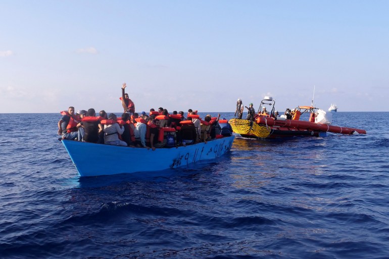 A small blue boat full of migrants and asylum seekers is approached from behind by a government vessel on the Mediterranean Sea. Each of the migrants wears an orange life vest. One of them stands up and raises an arm in the air to wave.