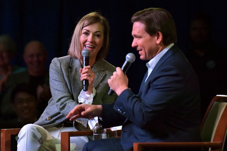 Kim Reynolds and Ron DeSantis sit side by side on a darkened stage, each holding a handheld microphone to their face as they speak.