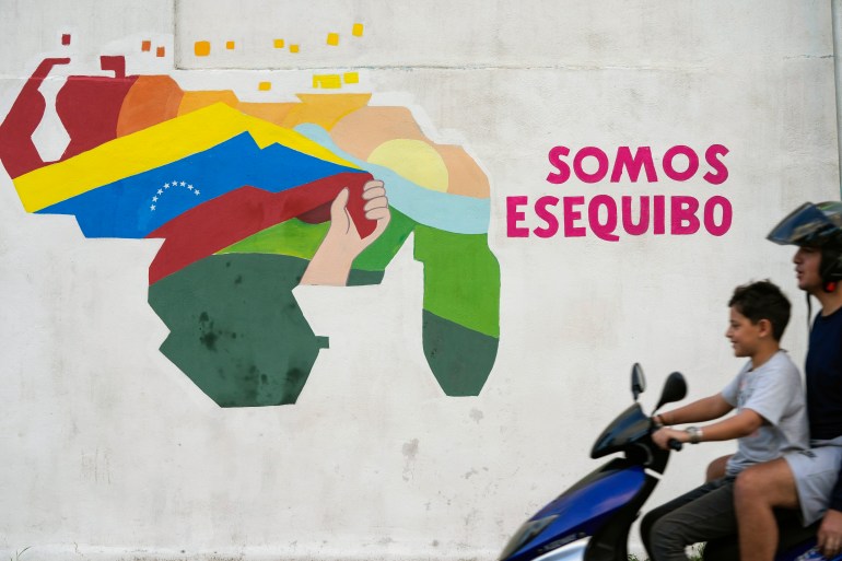 Dois meninos em uma motocicleta passam por um mural que mostra um mapa da Venezuela ampliado pela adição da região de Essequibo.  Dentro do mapa, uma mão agita a bandeira venezuelana.  Ao lado do mapa estão as palavras, 