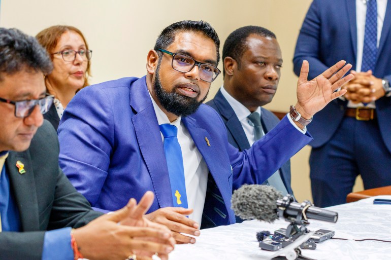 President Irfaan Ali, dressed in glasses and a bright blue suit with a white shirt and blue tie, speaks at a panel table, lifting one hand in the air to gesture.