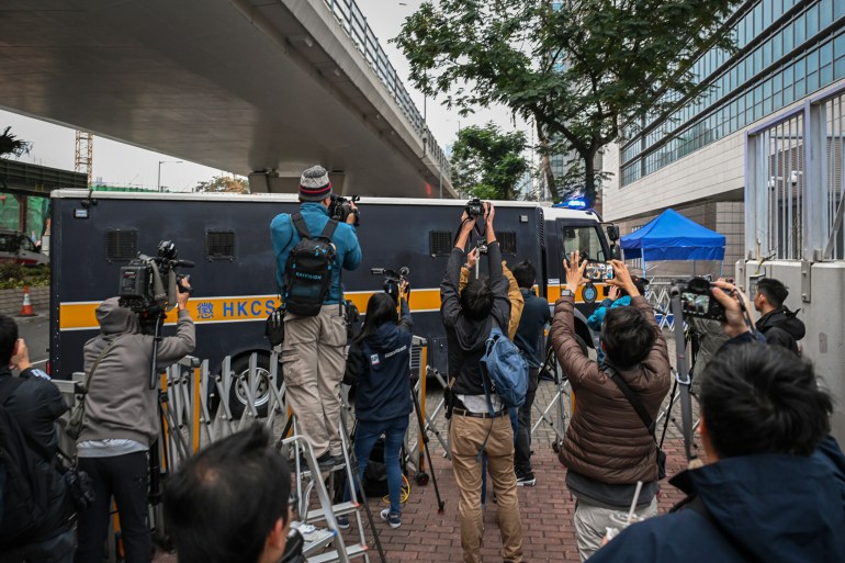 Journalists try to get a shot as the prison van carrying Jimmy Lai enters the court complex. They are standing on ladders and holding their cameras high. The van is black with a yellow stripe along the side.