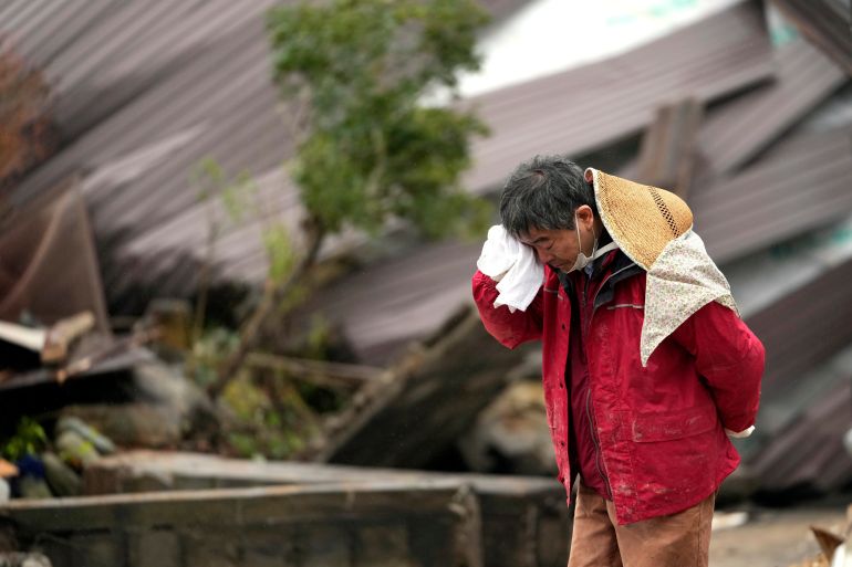 A man weeps as one of his family is found in their collapsed home. He is wearing a red jacket and is holding a white cloth to his face. The collapsed building is in the background