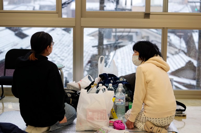 Due donne in un centro di evacuazione a Wajima.  Indossano abiti invernali e si siedono per terra.  La vista dalla finestra mostra i tetti innevati.