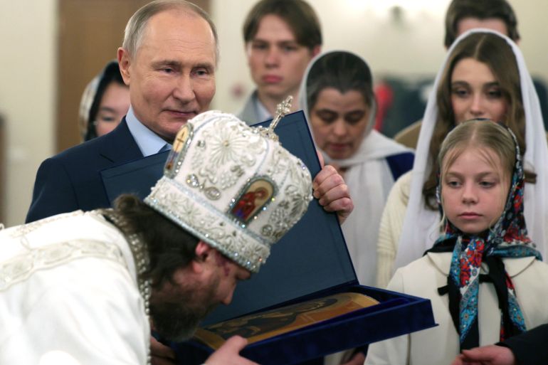 Putin smiling as he stands with the families of soldiers killed in Ukraine. A Russian Orthodox priest in white robes is kissing an icon in front of them.