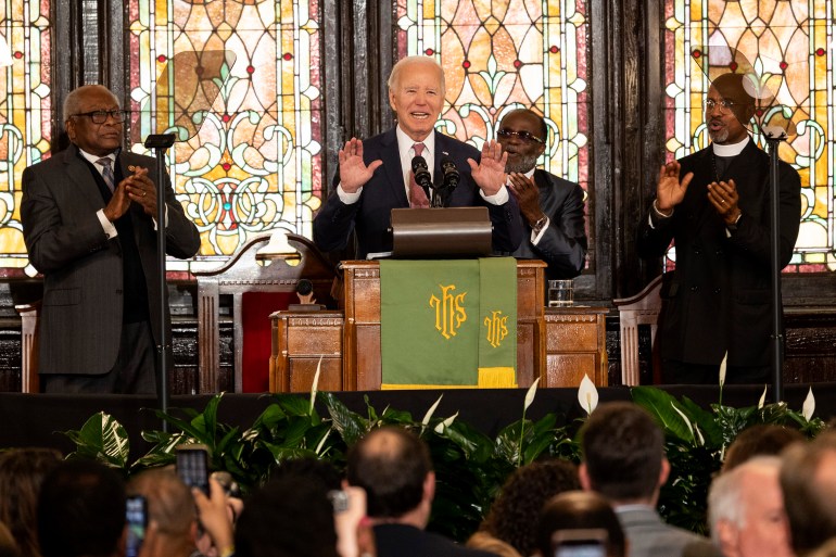 Joe Biden stands at the wooden pulpit of Mother Emanuel AME church in South Carolina, flanked by church leaders. Behind him are stained glass windows, and calla lilies and audience members sit in front of him.