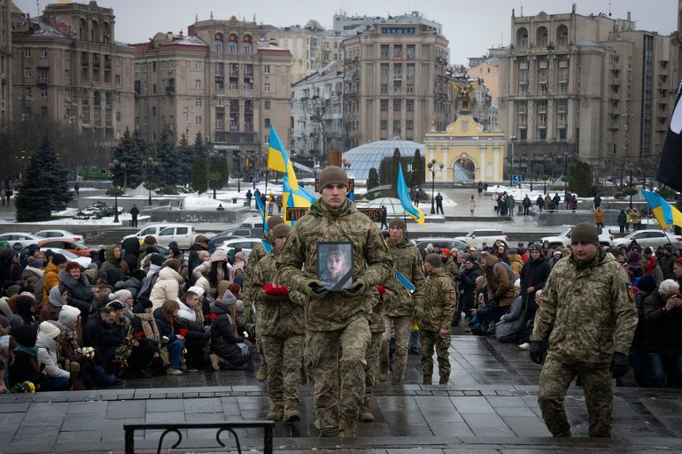 Ukraynalı asker ve şair Maksym Kryvtsov için cenaze töreni.  Savaş üniformalı bir asker, elinde bir fotoğrafla tabutunun önünde yürüyor.  Ukrayna bayrakları dalgalanıyor.  Etraftaki insanlar diz çöküyor.