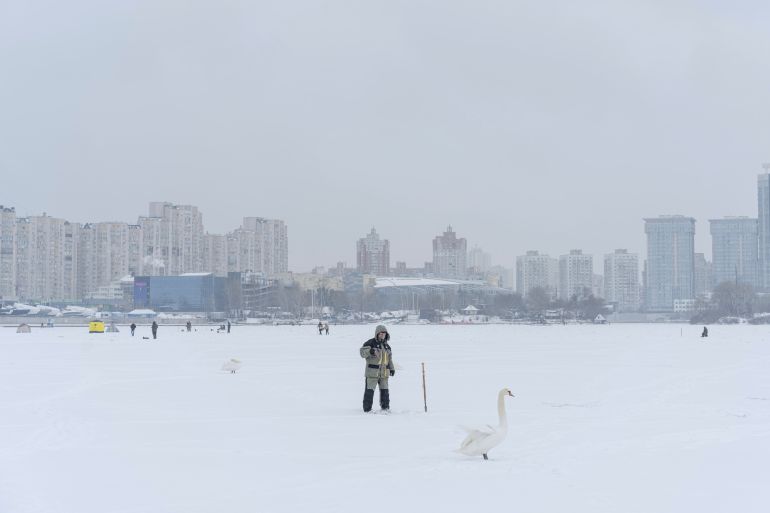 Un uomo che pesca attraverso un buco nel ghiaccio sul fiume Dnipro coperto di neve a Kiev.  Ci sono condomini dietro di lui e un cigno davanti.