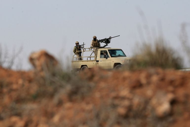 A military truck crosses the countryside near the Jordan-Syria border, with two soldiers standing in the truck bed, one attending a machine gun mounted on the cab's roof.