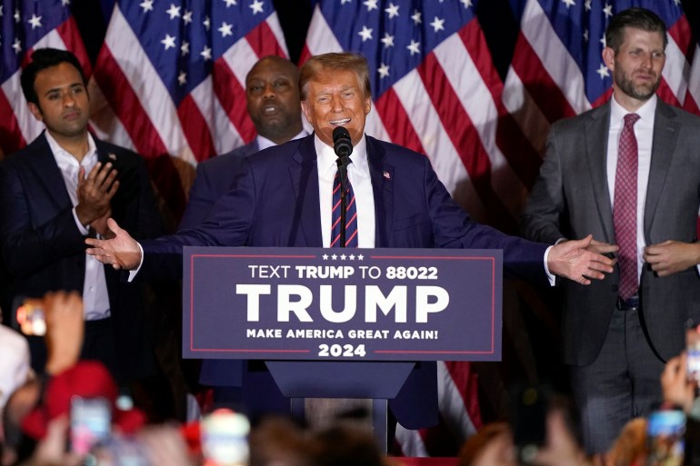 Donald Trump, adının yazılı olduğu podyumun arkasında kollarını iki yana açıyor.  Arkasında siyasi destekçiler ve bir dizi ABD bayrağı var.