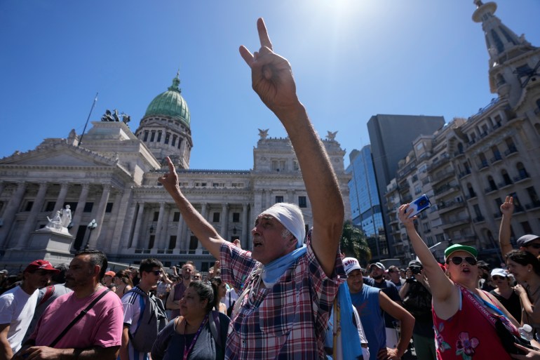 Un bărbat își ridică mâinile în aer, făcând semne de pace, în timp ce poartă steagul argentinian în fața clădirii Congresului din Buenos Aires.