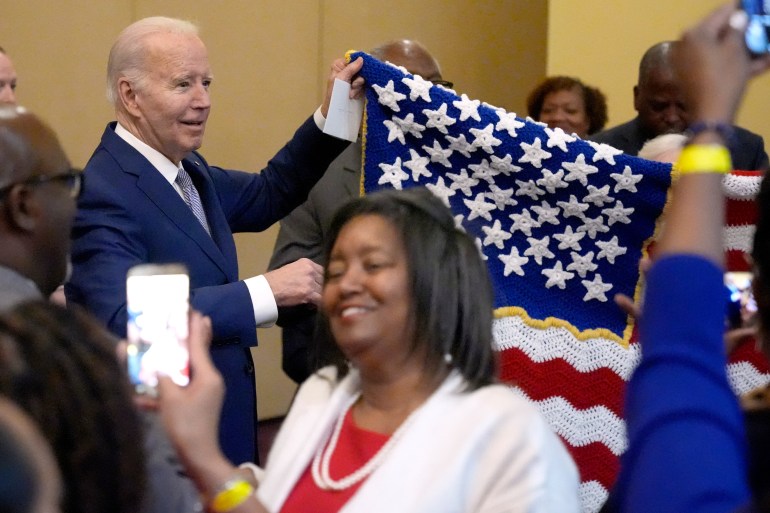 Joe Biden, dressed in a dark suit, holds up a crocheted US flag, while a woman poses for a cellphone photo.