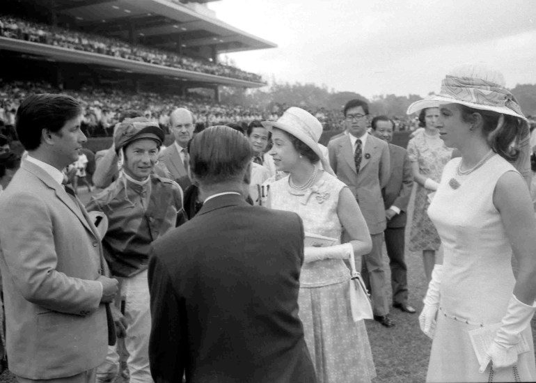 Queen Elizabeth Ii at the Singapore Turf Club in 1972. She is speaking to jockey Lester Piggott. Her daughter Princess Anne is with her. The grandstand is behind them. 