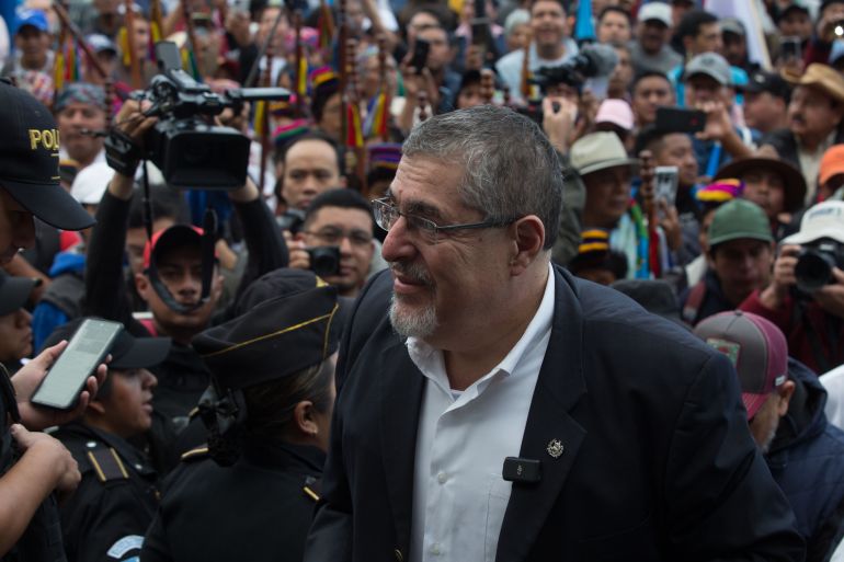 Bernardo Arevalo, dressed in a black blazer and white collared shirt, stands amid a crowd of supporters. One person behind him holds up a heavy video camera, similar to those used for TV broadcasts.