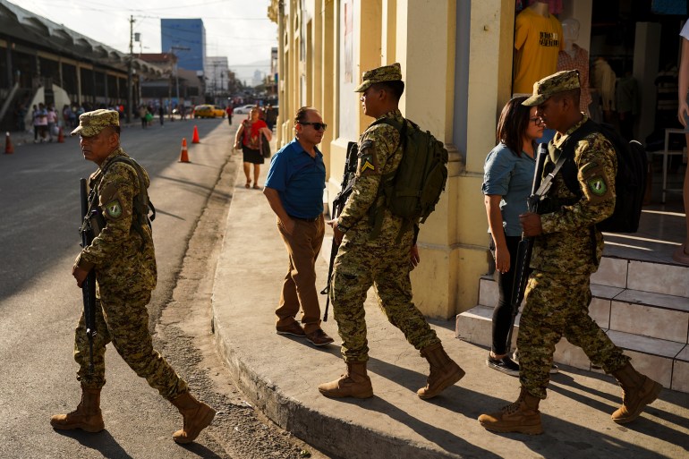Three soldiers, wearing beige fatigues and holding firearms, walk through the streets of San Salvador, past pedestrians.