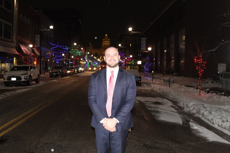 A smiling man in a dark blue suit and red tie stands on an Iowa road at nighttime, his hands folded. Snow lines the sides of the streets.
