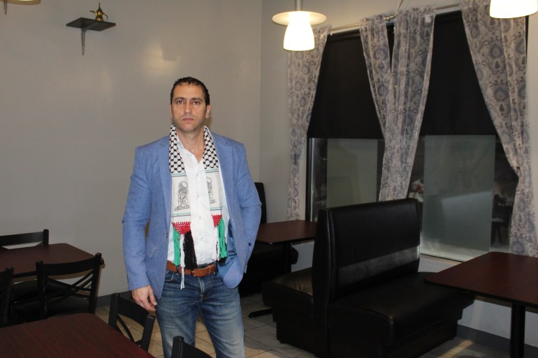A man stands in a room surrounded by windows, tables and chairs. He is dressed in a light blue blazer, jeans and a keffiyeh-style scarf that ends with a woven version of the Palestinian flag.