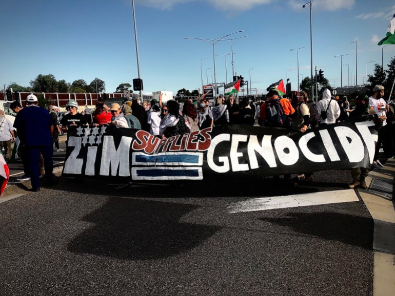 Manifestantes caminhando com uma faixa gigante dizendo ZIM=Genocídio.  Eles estão carregando a bandeira na frente deles enquanto avançam