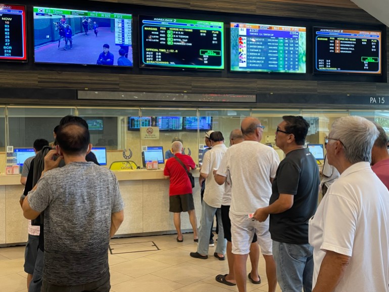 Men queue to place bets at the Singapore race course. They are dressed in T-shirts and shorts and are mostly older. There are screens above the counters.