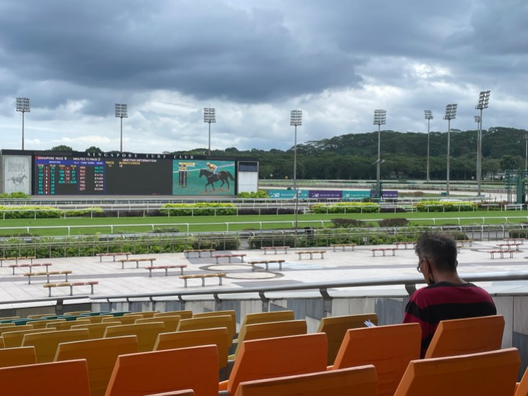 View of the racecourse in Singapore. There's one man in the stands. The board shows a race will start soon. It looks deserted