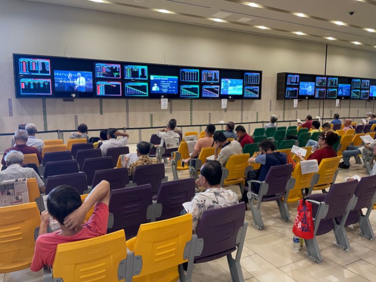 Spectators watch races from other parts of Asia during a lull in proceeedings. There are men sitting on seats with banks of screens against the wall at the front.
