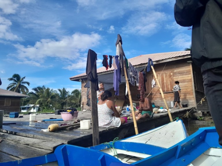 A wooden home sits perched on the water in Essequibo, surrounded by a dock and boats. Some Warao Indigenous people are seen sitting outside, preparing a meal under a clothesline. Palm trees are visible in the background.
