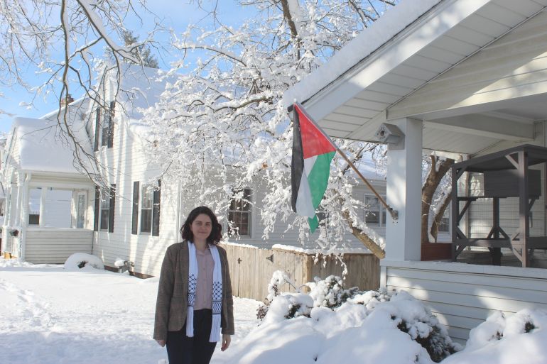 In a landscape blanketed white from heavy snowfall, a woman in a keffiyeh scarf and blazer stands next to a white house, the only colour on it coming from a bright Palestinian flag.