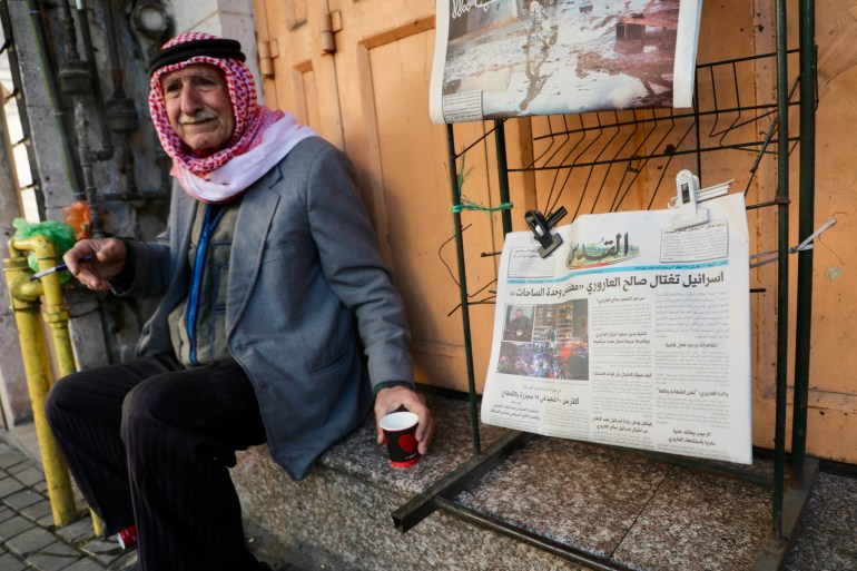 Newspaper seller on a shuttered shop's stoop, headline reads "Israel assassinated Saleh al-Arouri"