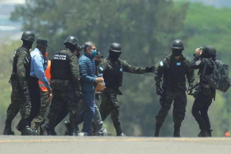 Honduras national police, dressed in helmets and flak jackets, escort former President Juan Orlando Hernandez across a road.