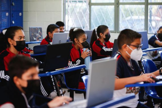 Students wearing COVID-19 face masks sit behind their desks and type at their laptops. One slips a water bottle under her mask to drink.