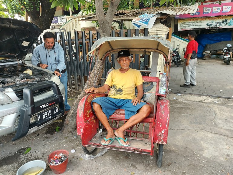 Sarti sitting in his pedicab. It is parked under a tree on the side of the road. He is wearing a T-shirt, shorts and flip flops. Another man is working on a car next to him and the bonnet is up.