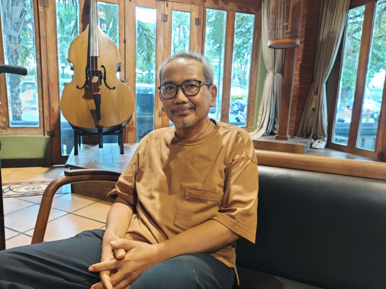 Slamet Raharjo. He is seated on a wooden framed seat inside his home. There is. double bass behind him near the window.