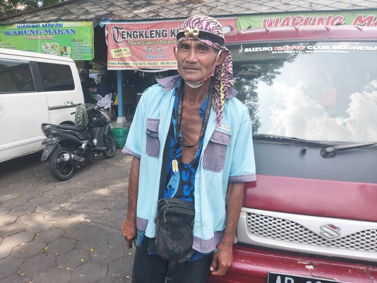 Car park attendant Tawar. He is leaning against a van. He has a cloth tied around his head and a is wearing long trousers, a shirt open nearly to his waist and a uniform shirt over that. 