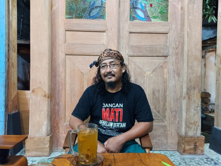 Management consultant Indrawan, He is seated in front of a wooden door and smiling. He has a large glass of tea in front of him.