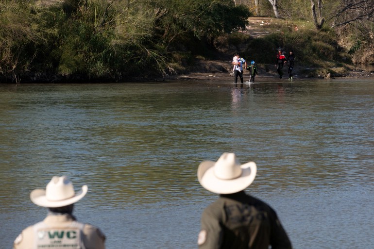 Two men in ten-gallon hats watch migrants wade into the Rio Grande.