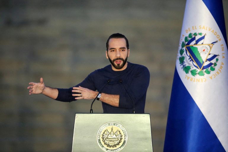 Nayib Bukele, dressed in a black long-sleeve shirt, stands behind a podium and gives a speech with his arms outstretched in gesture. Behind him is a Salvadoran flag.