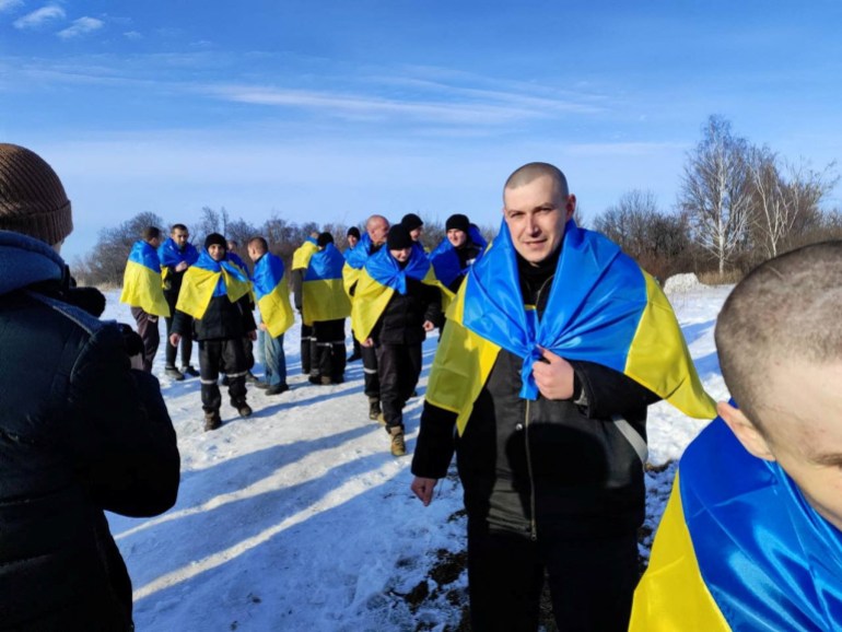 Ukrainian prisoners of war walk across a snow-covered field, each person wearing a Ukrainian flag across their shoulders like a cape.