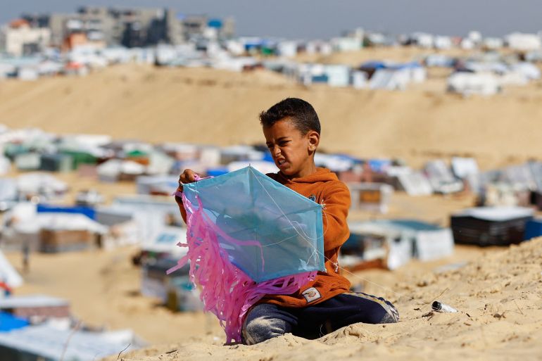 A small boy builds a kite against the background of tents and desperation.