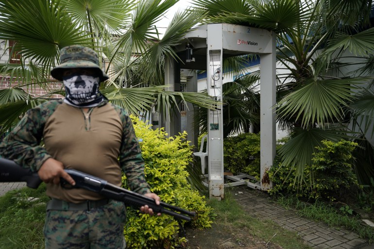 Um militar armado, vestido com camuflagem, capacete, máscara facial e colete, fica em frente a um scanner corporal cercado por palmeiras e arbustos.  Um prédio de tijolos pode ser visto através da vegetação.