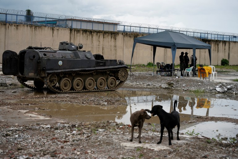 Um tanque fica do lado de fora do muro de uma prisão em Guayaquil.  O chão está lamacento e, em primeiro plano, dois cachorros farejam um ao outro.  Uma barraca de dossel com algumas pessoas embaixo pode ser vista ao fundo.