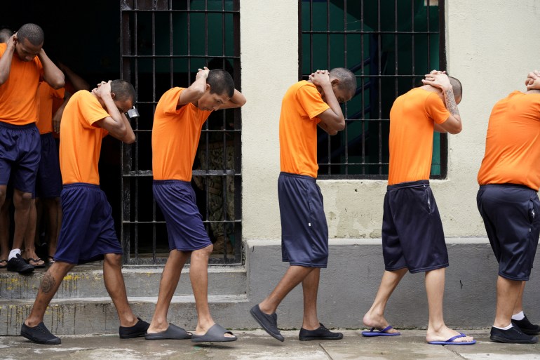 Prisioneiros com camisas laranja e shorts andam em fila única, com as mãos atrás da cabeça voltada para baixo.