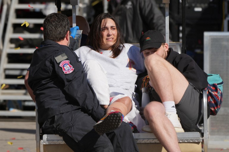 A first responder looks at an IV drip for a woman whose leg is propped up on his leg. She wears a white sweatshirt and appears to be in pain. She sits on a bench with a man in a ballcap.
