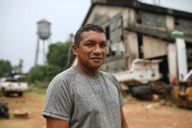 A man in a grey T-shirt stands in front of the abandoned warehouses at Fordlandia, an industrial town in the Amazon. A water tower is also visible behind him.