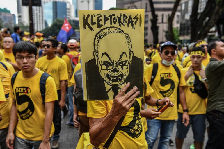 Protesters marching in Malaysia over the 1MDB scandal. They are wearing yellow T-shirts, One is carrying placard showing a caricature of Najib beneath the words 'bapak kleptokrasi' (father of kleptocracy)