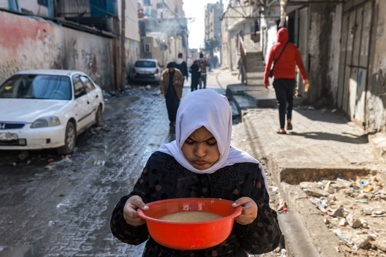 Uma garota carregando uma tigela de sopa de lentilhas.  Ela está andando pelas ruas de Rafah.  Ela está usando um vestido preto com um lenço branco na cabeça.