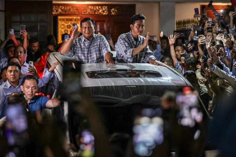 Prabowo e Gibran in un'auto scoperta salutano i sostenitori dopo che un rapido conteggio dei voti ha mostrato che hanno vinto le elezioni del 2024. C'è folla intorno all'auto circondata dalla sicurezza.  Prabowo saluta.  Onde di Gibran