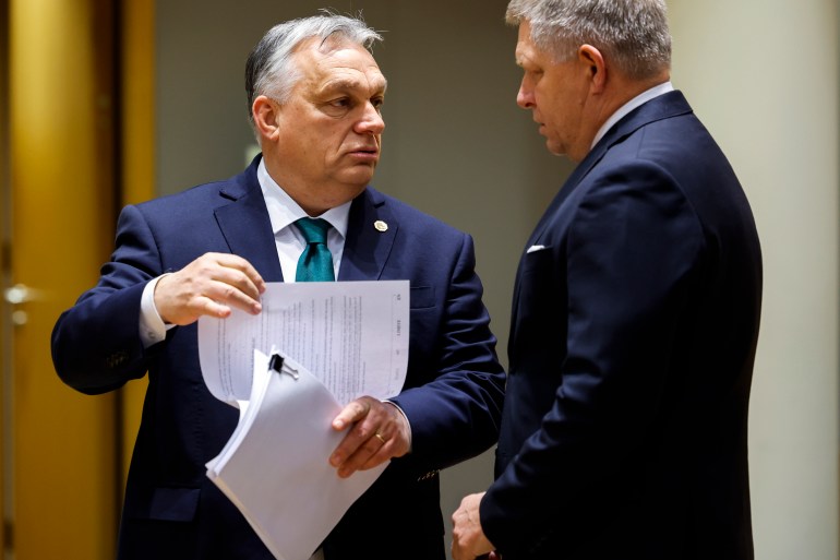 Victor Orban segura um pacote de papéis, levantando uma folha enquanto fala com Robert Fico.  Ambos os homens usam terno e gravata.